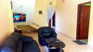 Una sala de estar con un sofá negro, un sillón negro y una pequeña mesa de centro. Hay un televisor en la pared y una escalera al fondo.