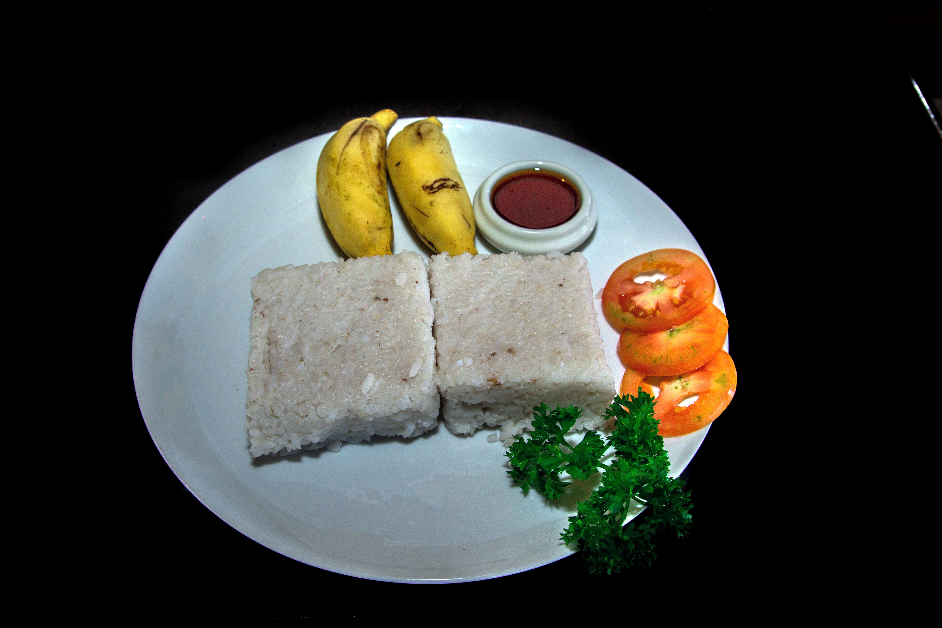 Un plato con dos porciones de comida blanca, dos bananas, un tomate y un recipiente pequeño con salsa.