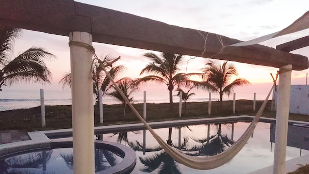 A pool area with a hammock, palm trees, and a view of the ocean at sunset. - La Casa de la Playa (Cuyutlan)