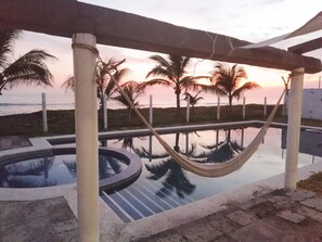 A pool area with a hammock, palm trees, and a view of the ocean at sunset.