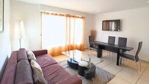 A modern living room with a sofa, a glass-top coffee table, a dining table with chairs, and a flat-screen TV mounted on the wall. - Viva Riviera 2 Bedrooms 2 Bathrooms Rue Florian (Cannes)