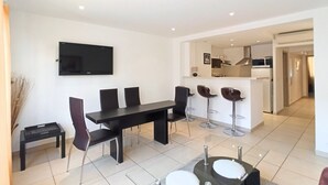 A modern living room with a dining area, a kitchen in the background, and a TV mounted on the wall. - Viva Riviera 2 Bedrooms 2 Bathrooms Rue Florian (Cannes)