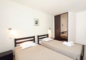 Quarto de hotel com duas camas de solteiro, uma pequena mesa de cabeceira, um criado-mudo, um espelho fixado na parede e um quadro emoldurado.