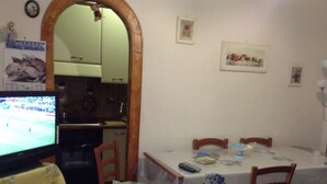 A dining area with a table set for two, a television on the left, and a kitchen visible through an archway. - MOUNTAIN HOUSE PESCASSEROLI (pescasseroli)