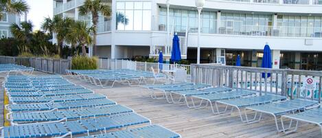 A poolside area with blue lounge chairs and umbrellas, a wooden deck, and a white building with balconies.