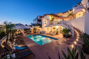 A pool area with lounge chairs and umbrellas, a staircase, and a building with balconies and plants.