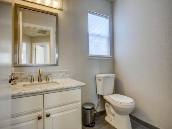 A bathroom with a white toilet, a sink with a marble countertop, a mirror above the sink, and a window with blinds.