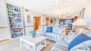 A living room with a striped sofa, a white coffee table, and a bookshelf filled with books. - Semi-Oceanfront Condo w/ Private Balcony, Resort Pool, Elevator, Beach Access (Rodanthe)