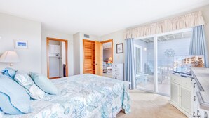 A bedroom with a bed, a dresser, a desk, and a view of a pool and buildings through a glass door. - Semi-Oceanfront Condo w/ Private Balcony, Resort Pool, Elevator, Beach Access (Rodanthe)