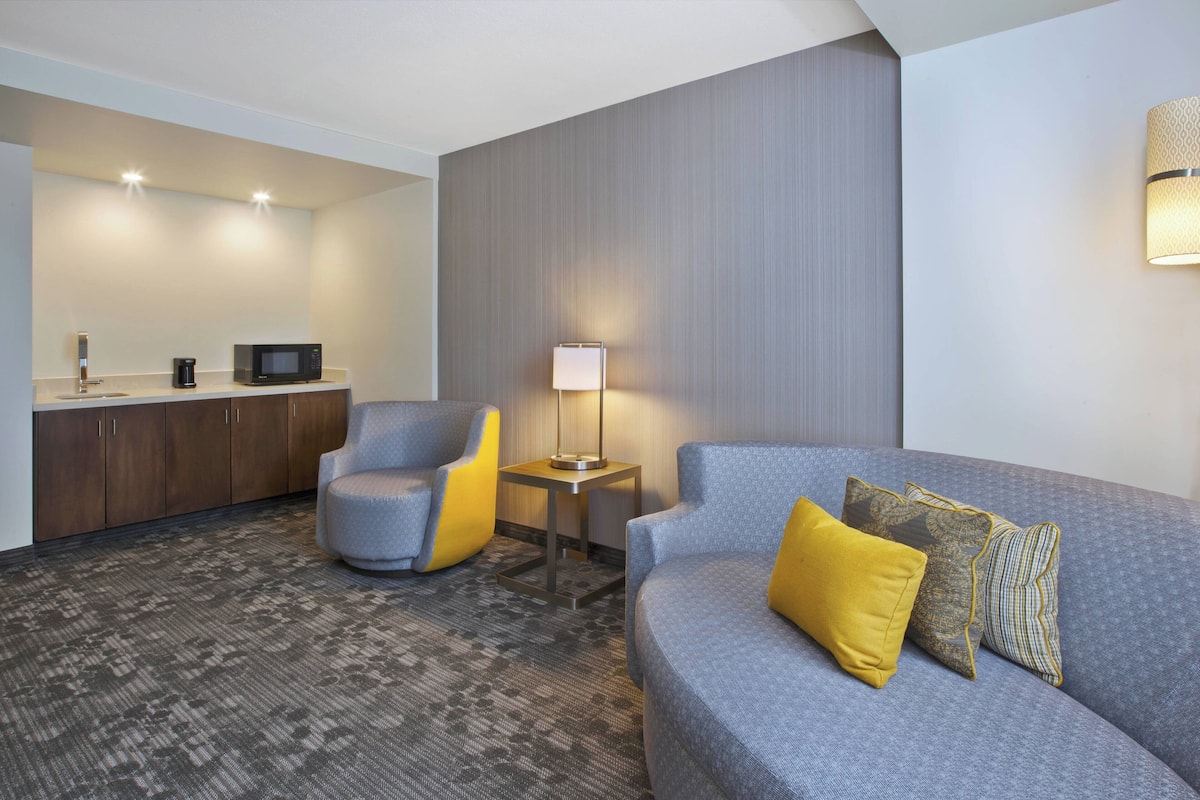 Een hotelkamer met een grijze bank, een gele fauteuil, een tafeltje met een lamp, een magnetron en een koffiezetapparaat.