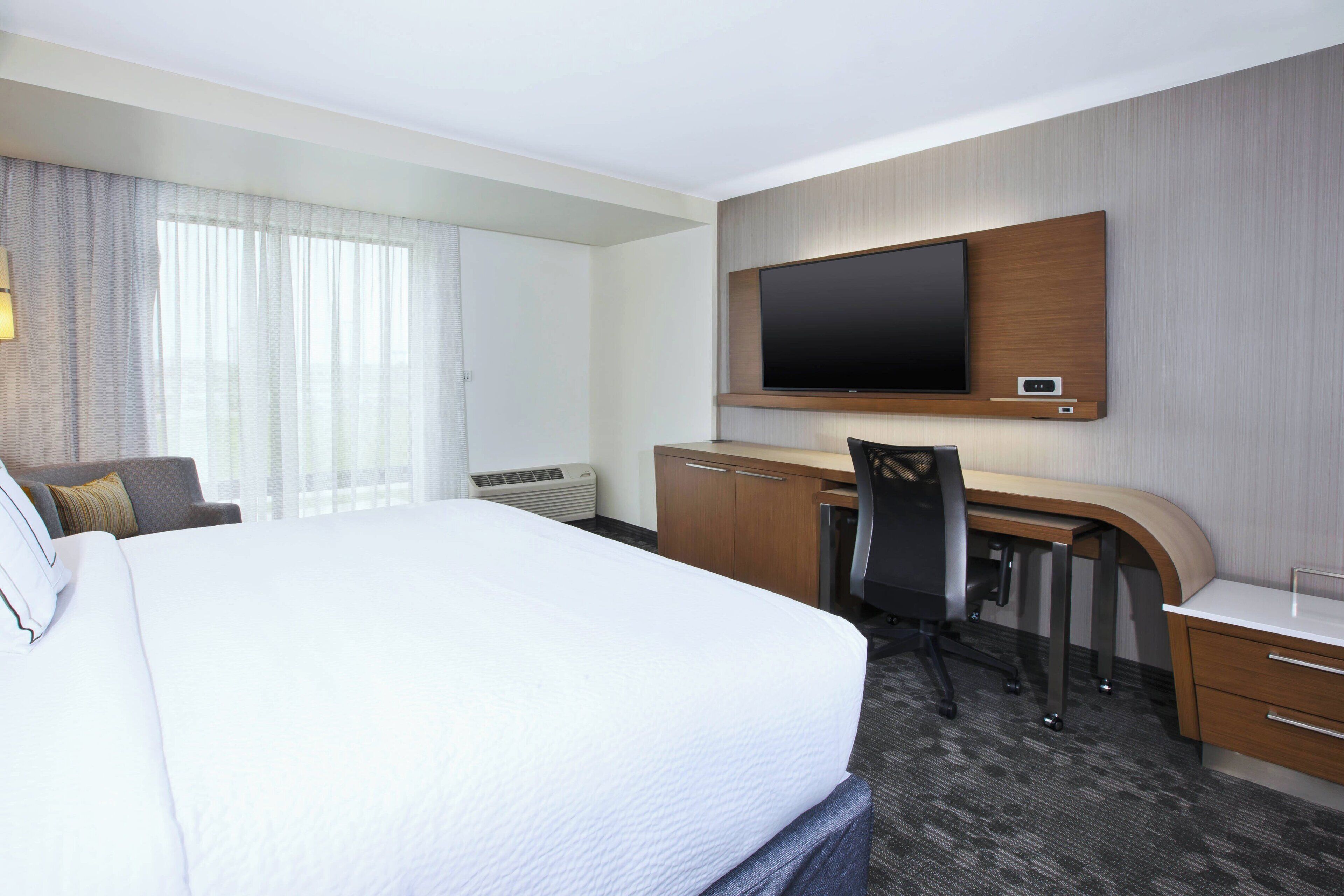 A hotel room with a large bed, a desk with a chair, a flat-screen TV mounted on the wall, and a window with curtains.