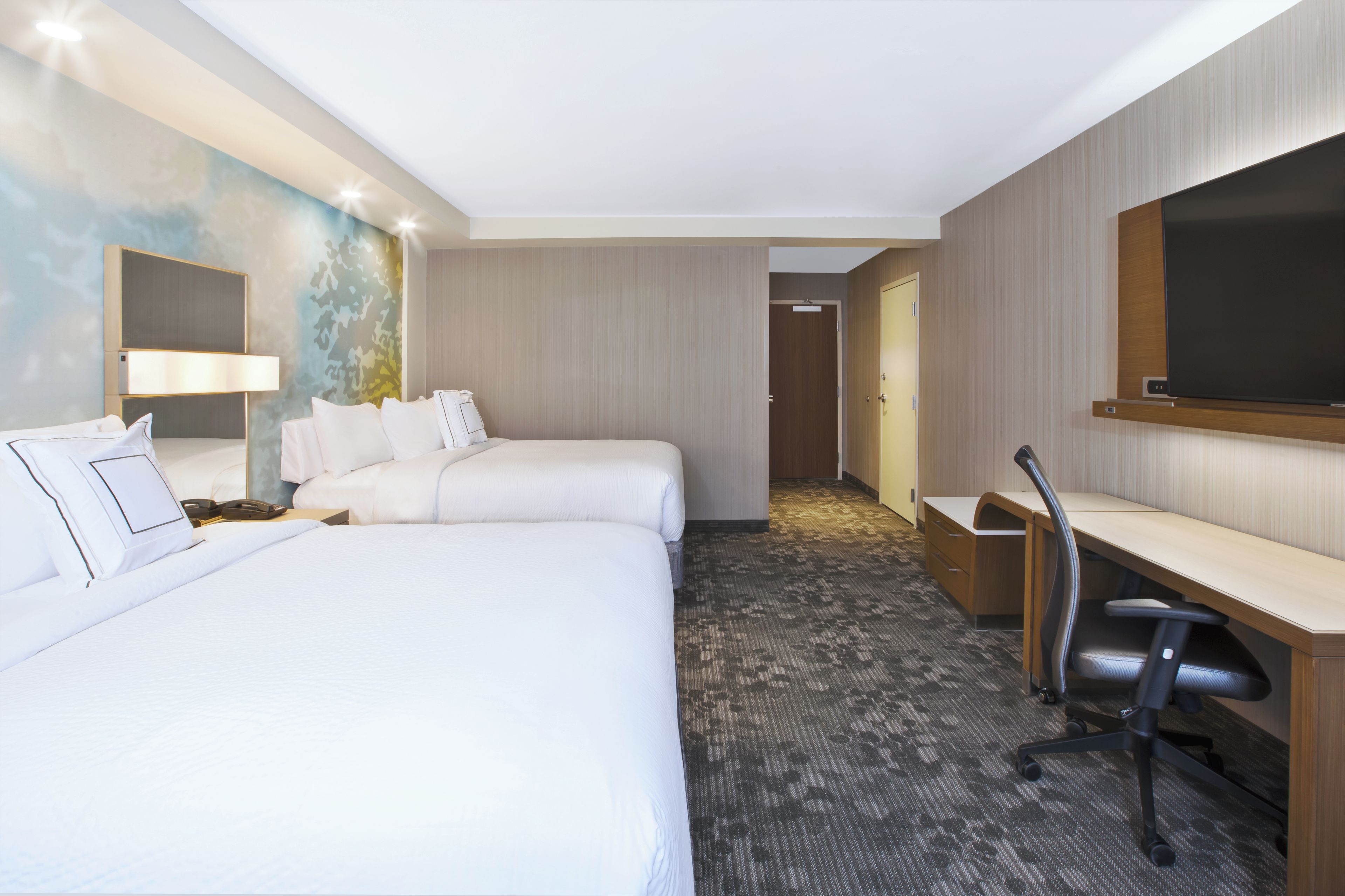 A hotel room with two beds, a desk, a chair, a television, and a bathroom visible through an open door.