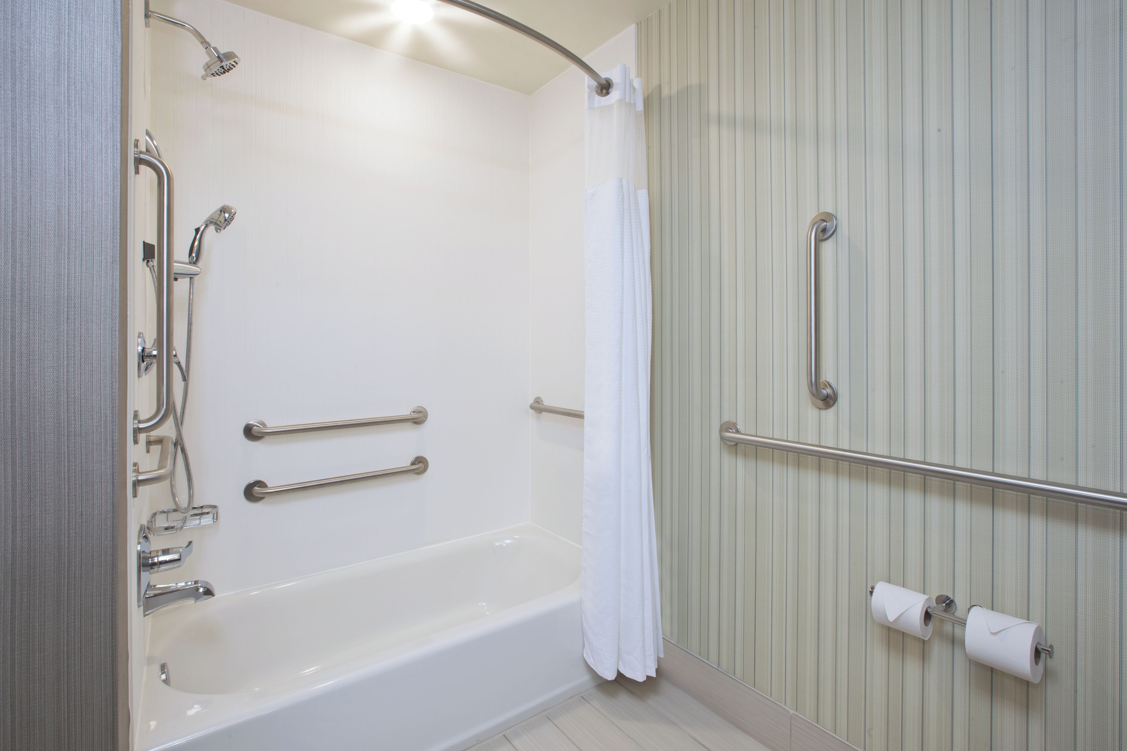 A bathroom with a bathtub, grab bars, a showerhead, and a toilet paper holder.
