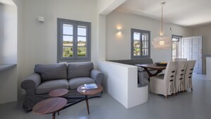 A modern living room with a grey sofa, two round wooden tables, and a dining area with a wooden table and four chairs. - Villa Anemos (Ampelas)