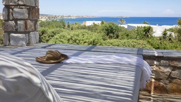 A bed with a straw hat, stone pillar, and a view of the sea and buildings. - Villa Anemos (Ampelas)