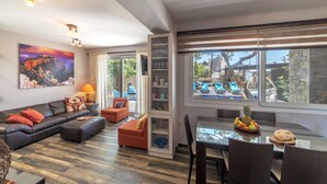 Un salon moderne avec un canapé, un pouf et une table à manger avec des chaises. Une grande fenêtre offre une vue sur l’extérieur.