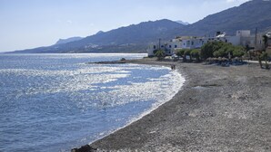 Une plage de galets, une mer d’un bleu limpide et des bâtiments en arrière-plan.