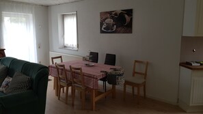 A dining area with a table set for four, surrounded by wooden chairs. A window with sheer curtains allows natural light to enter. A painting hangs on the wall above the table. - Apartment in a quiet location (Selters (Taunus))