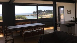 A room with a large window overlooking a beach, a dining table with chairs, and a kitchen area. - Waterfront house Piriac sur mer (Piriac sur mer)