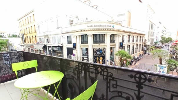 Ein Balkon mit grünen Stühlen und einem Tisch mit Blick auf eine Straße mit Gebäuden und einem Schild für 'Société Marseillaise de Crédit'.