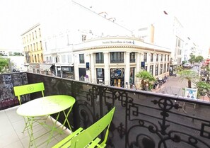 Un balcon avec des chaises vertes et une table, donnant sur une rue bordée d’immeubles et sur laquelle on peut voir une enseigne indiquant « Société Marseillaise de Crédit ».