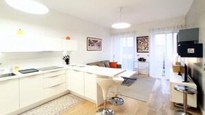 Eine moderne Küche mit weißen Schränken, einer zentralen Kochinsel mit Barhocker, einem Essbereich mit einer Couch und einem Fernseher im Wohnzimmer.