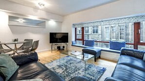 Ett modernt vardagsrum med en TV, lädersoffor, ett soffbord i glas och ett stort fönster med utsikt över byggnader.