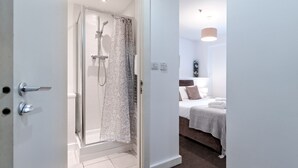 Ett hotellrum med dusch, en säng med ett vitt och grått randigt sängöverkast och ett litet nattduksbord med en lampa.