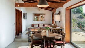 A dining area with a wooden table and chairs, a hanging light fixture, and a view of the outdoors through a large window.
