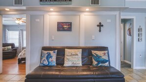 Una sala de estar con un sofá de cuero negro, dos cojines decorativos, un letrero en la pared que dice "Beach House" y una pequeña cruz decorativa.