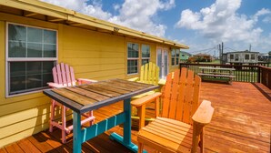 Una terraza de madera con una mesa y sillas, un edificio amarillo con ventanas, y un cielo azul despejado con nubes.