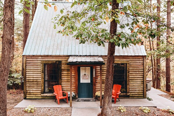 Une cabane rustique avec un toit en métal, des murs en bois et une porte d ’entrée. On peut voir deux chaises rouges à l’extérieur.