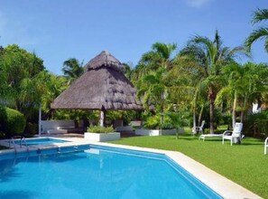 A swimming pool with a thatched-roof structure, surrounded by palm trees and greenery. - Large House-walking to beach-10 guests! (Zona Hotelera)