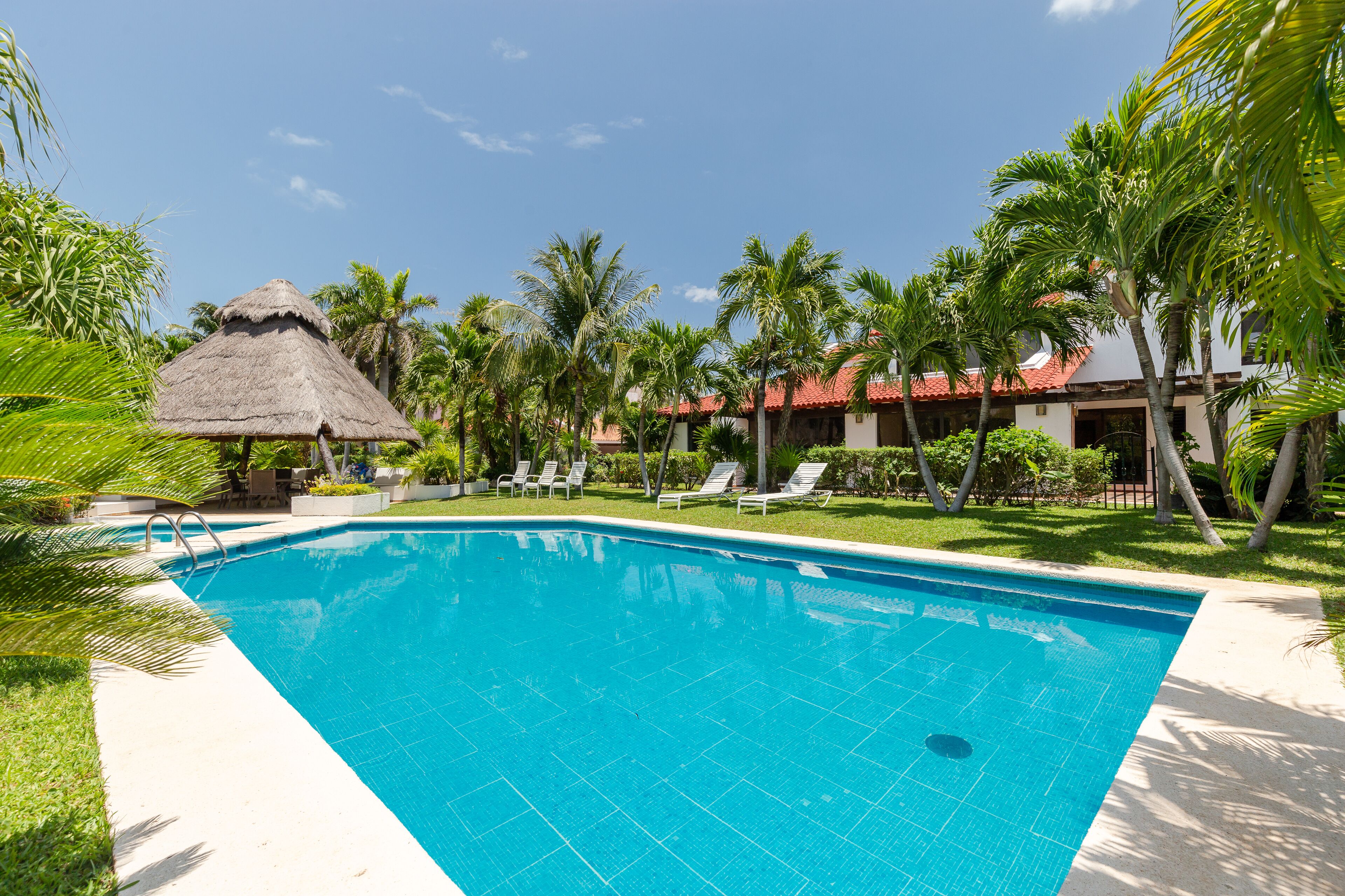 Una piscina azul cristalina rodeada de palmeras y sillas de descanso, con una estructura de techo de paja cerca.