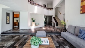 Un amplio salón con un sofá gris, una mesa de centro de madera con una planta y una escalera que sube a un nivel superior.