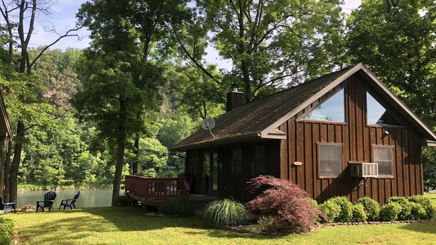 Une cabane dotée d’une terrasse, entourée d’une végétation luxuriante et bordée par un lac.