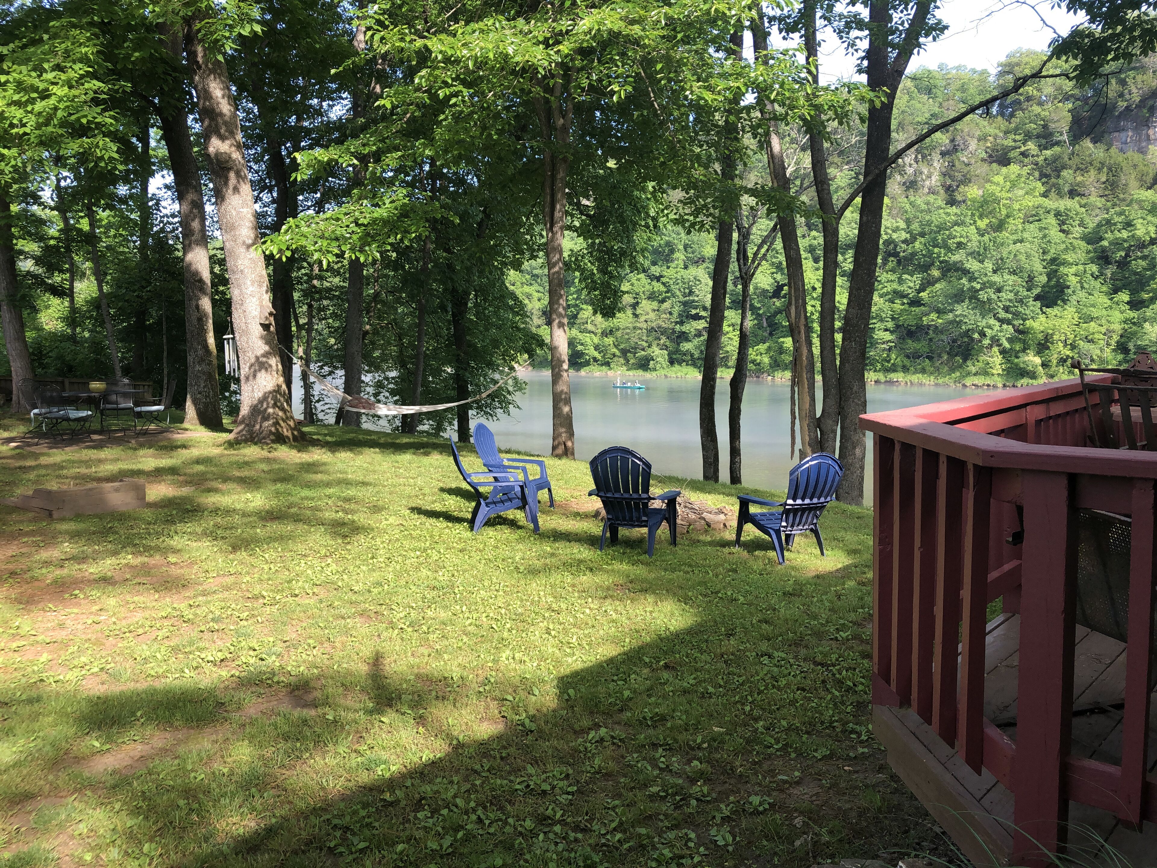 A lakeside deck with Adirondack chairs and a hammock overlooking a calm lake, surrounded by dense trees.