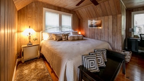 Een gezellige slaapkamer met een houten plafond, een bed met veel kussens, een nachtkastje met een lamp en een bureau met een stoel.