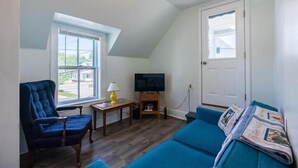 A small, cozy room with a sloped ceiling, a television, a blue armchair, a blue sofa, and a window with a view of the outside. - Ocean Breezes at Salty Air Suites, Suite C – New Balcony, 3BR, walk to the Beach (Old Orchard Beach)
