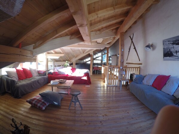A spacious loft-style room with high ceilings and wooden beams, featuring a large sofa, a smaller couch, and a dining table.