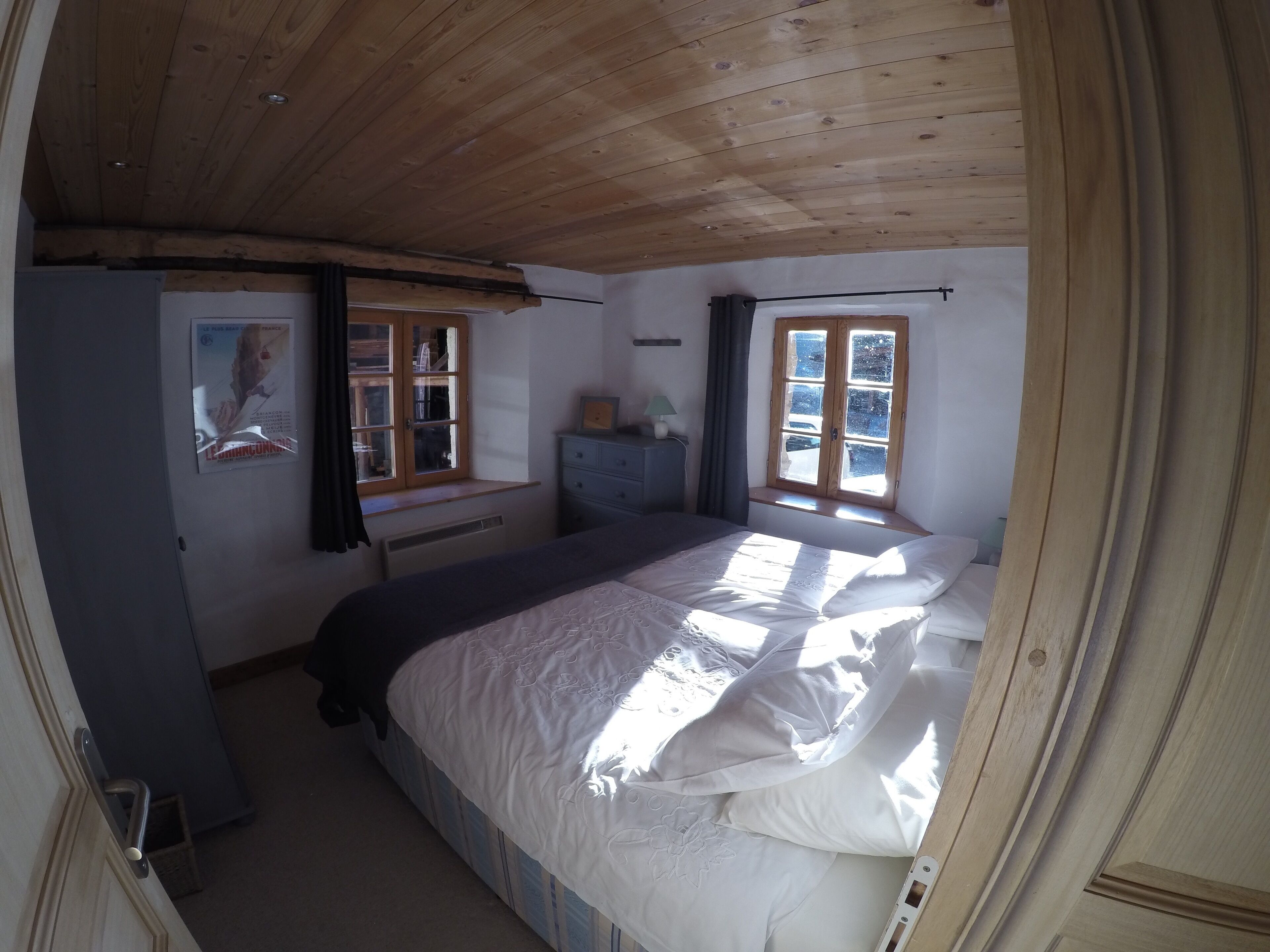 Un dormitorio con techo de madera, una cama con ropa de cama blanca, una ventana con cortinas y un aparador.