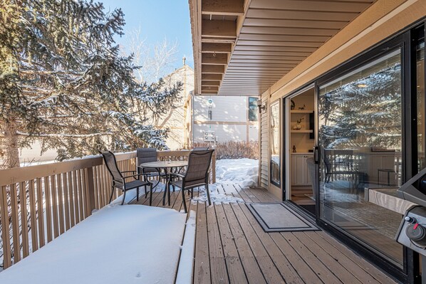 Une terrasse en bois avec une table et des chaises, une porte vitrée donnant sur une pièce et un sol recouvert de neige.