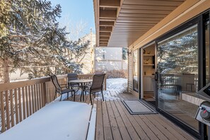 Um deck de madeira com mesa e cadeiras, uma porta de vidro que dá para um quarto, e chão coberto de neve.