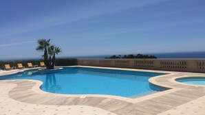 En pool med buet design, omgivet af liggestole og palmer, med udsigt over havet og en klar himmel.