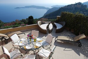 An outdoor dining area with a table set for four, surrounded by wicker chairs and a lounge chair, overlooking a scenic view of the sea and hills. - Stunning penthouse with panoramic views of Eze & Cap Ferrat (Èze, Provence-Alpes-Côte-D’Azur)