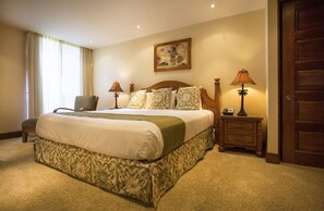 Un dormitorio de hotel con una cama grande, una silla, dos mesitas de noche con lámparas, un cuadro en la pared y una puerta de madera.