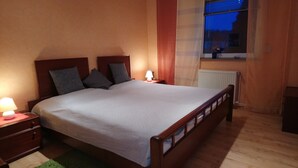 A neatly made double bed with a wooden frame, two bedside tables with lamps, a window with a view of the outside, and a radiator on the floor.