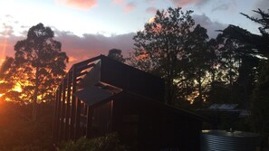 Un bâtiment moderne doté d’un toit en pente et de grandes fenêtres, entouré d’arbres, avec un coucher de soleil en arrière-plan.