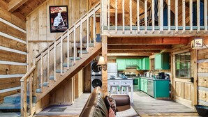 Interior de una cabaña de madera con escalera, sofá, zona de cocina y mesa de comedor.
