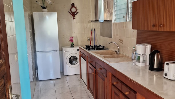 Une cuisine compacte équipée d’un lave-linge, d’un réfrigérateur, de meubles en bois, d’un évier et d’une fenêtre située au-dessus du plan de travail.